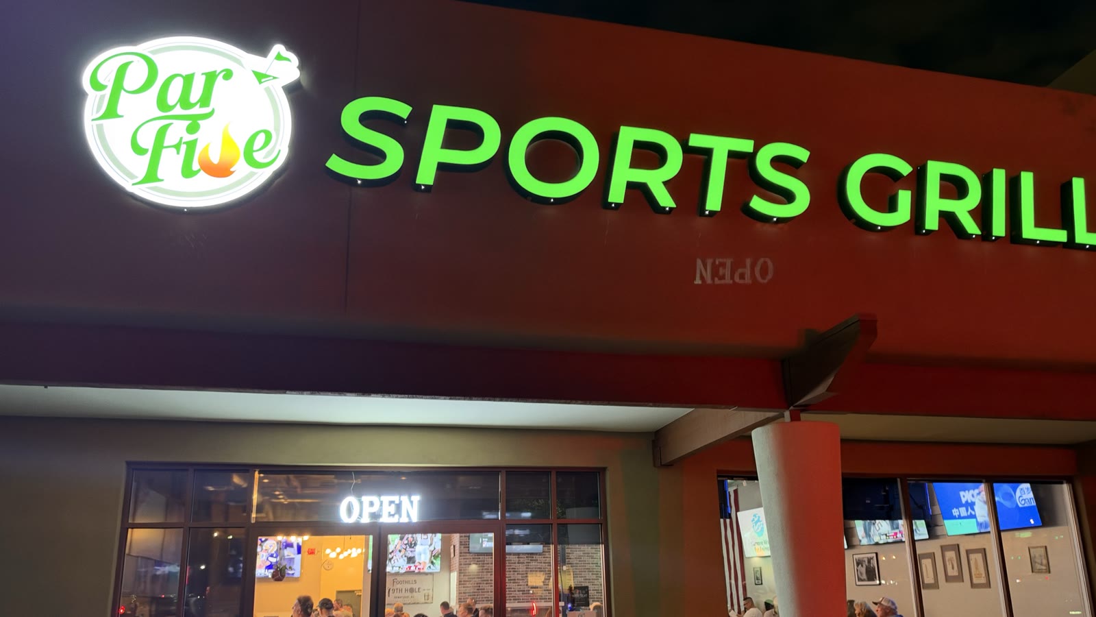 Par Five Sports Grill's lit storefront at night, with an Open sign on the door and diners inside